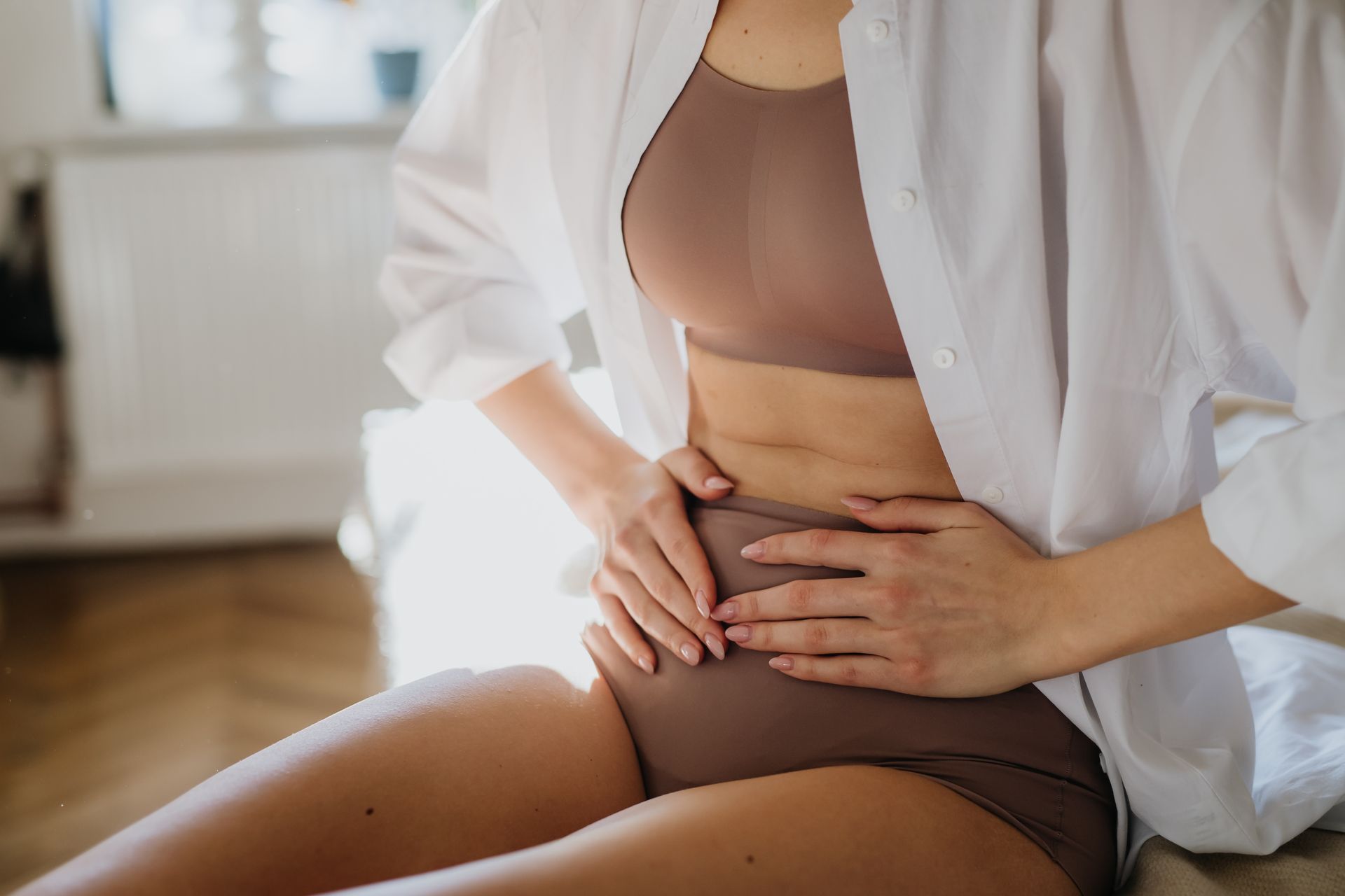 Woman in underwear and open white shirt holds her abdomen, likely experiencing pain.