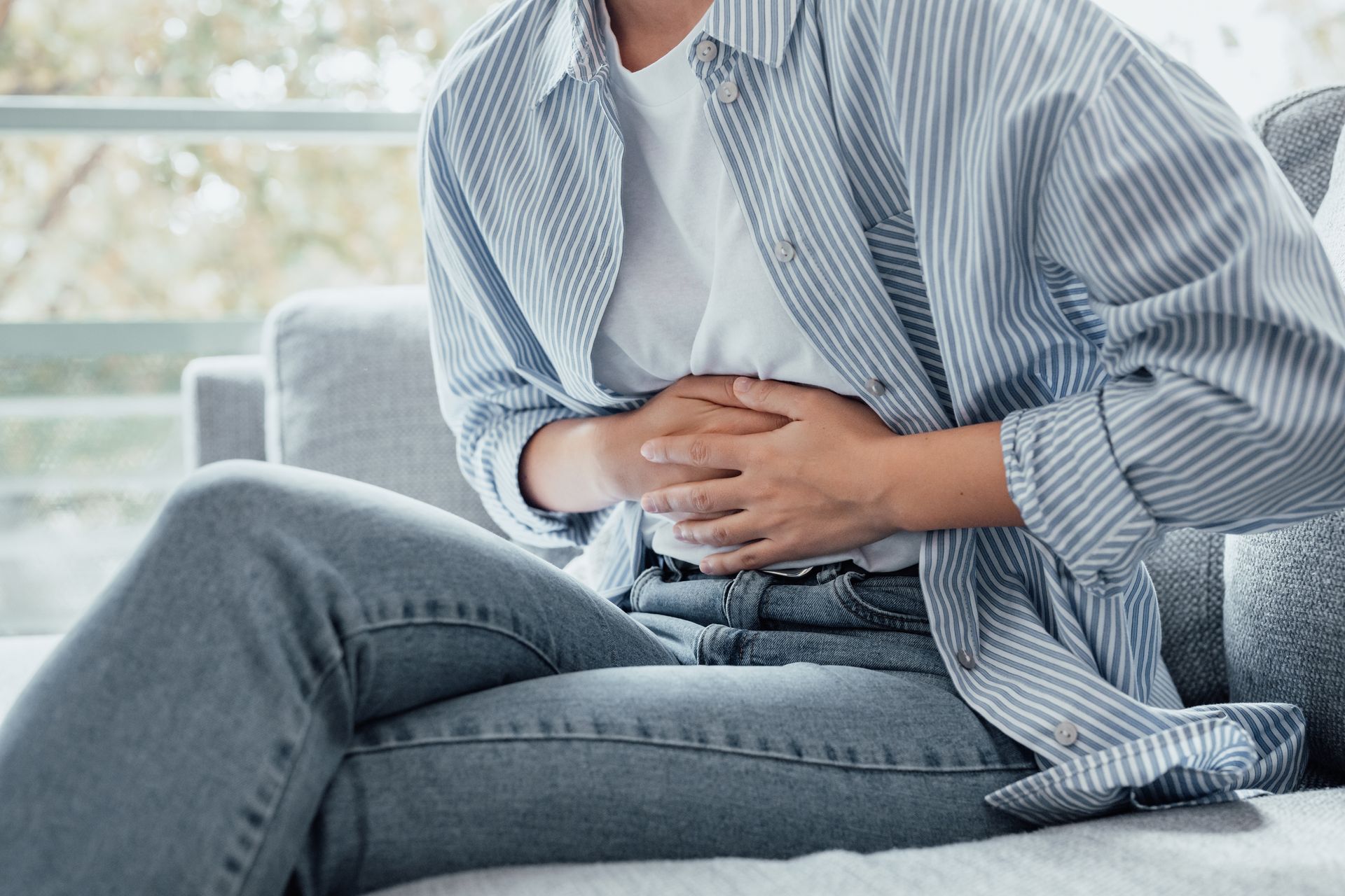 Woman holding stomach, possibly in pain, sitting on a gray couch, wearing jeans and a blue striped shirt.