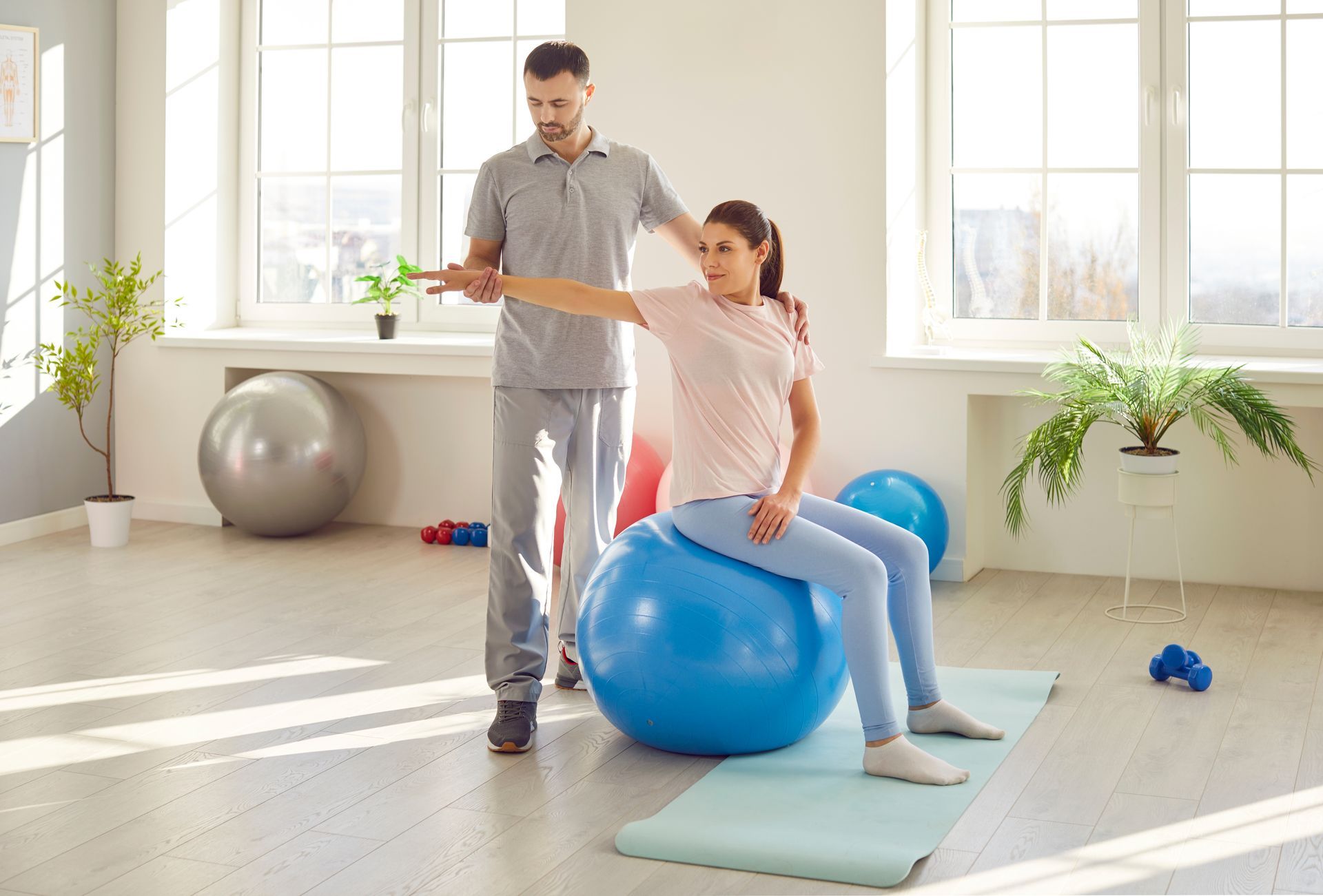 A woman is sitting on a blue exercise ball while a man helps her.