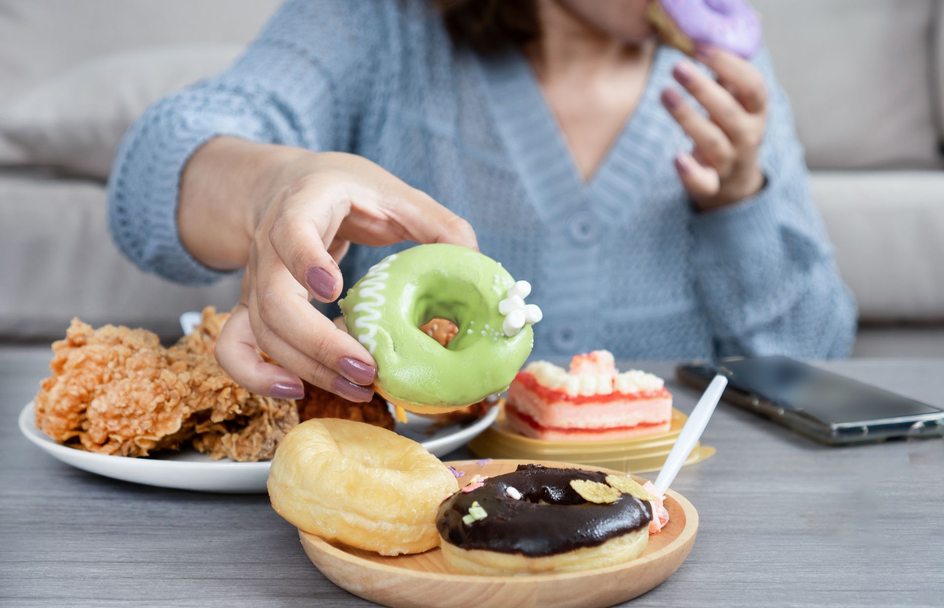 Woman reaching for green donut, surrounded by fried chicken, cakes, and other sweets.