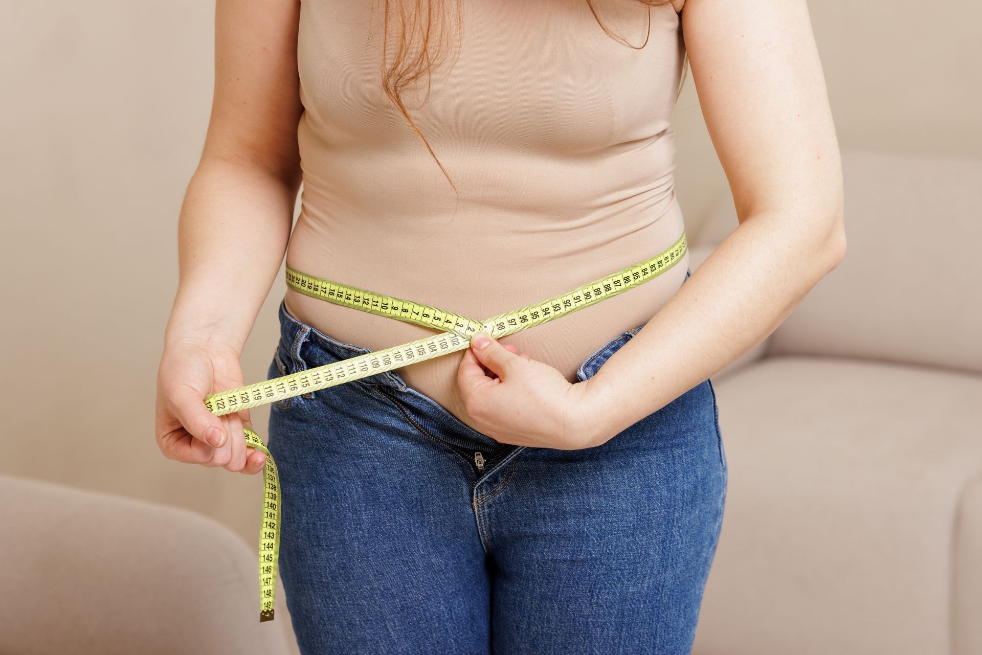 Woman in jeans and tank top measures her waist with a yellow tape measure indoors.