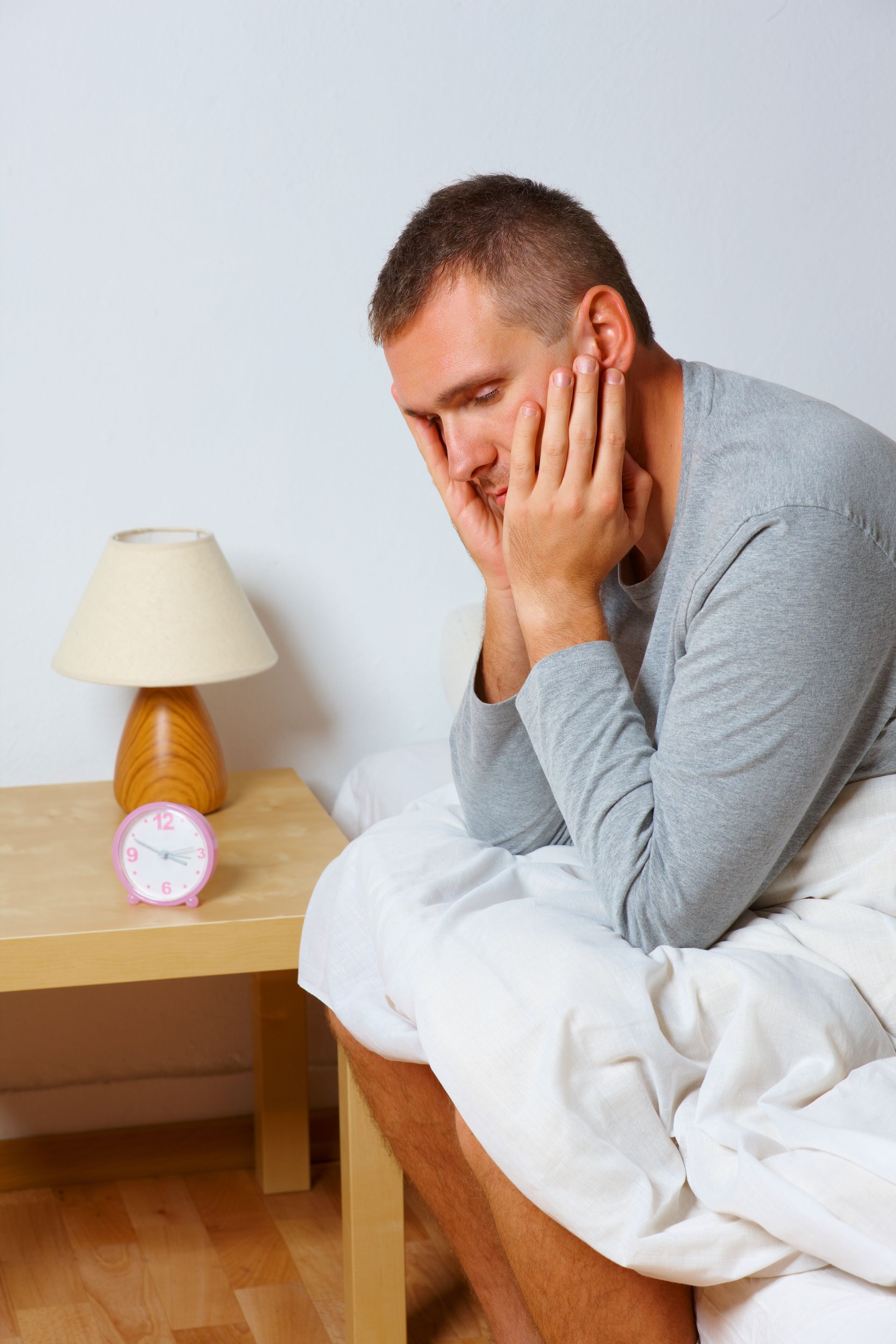 Man sitting on bed, looking tired, hands on his face. Alarm clock and lamp on nightstand.