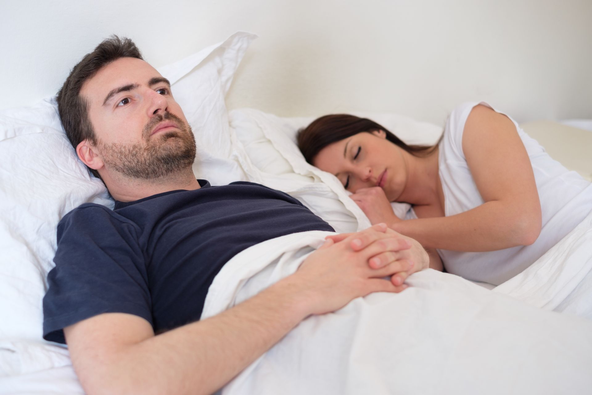 Man awake in bed, looking concerned. Woman asleep beside him. White sheets.