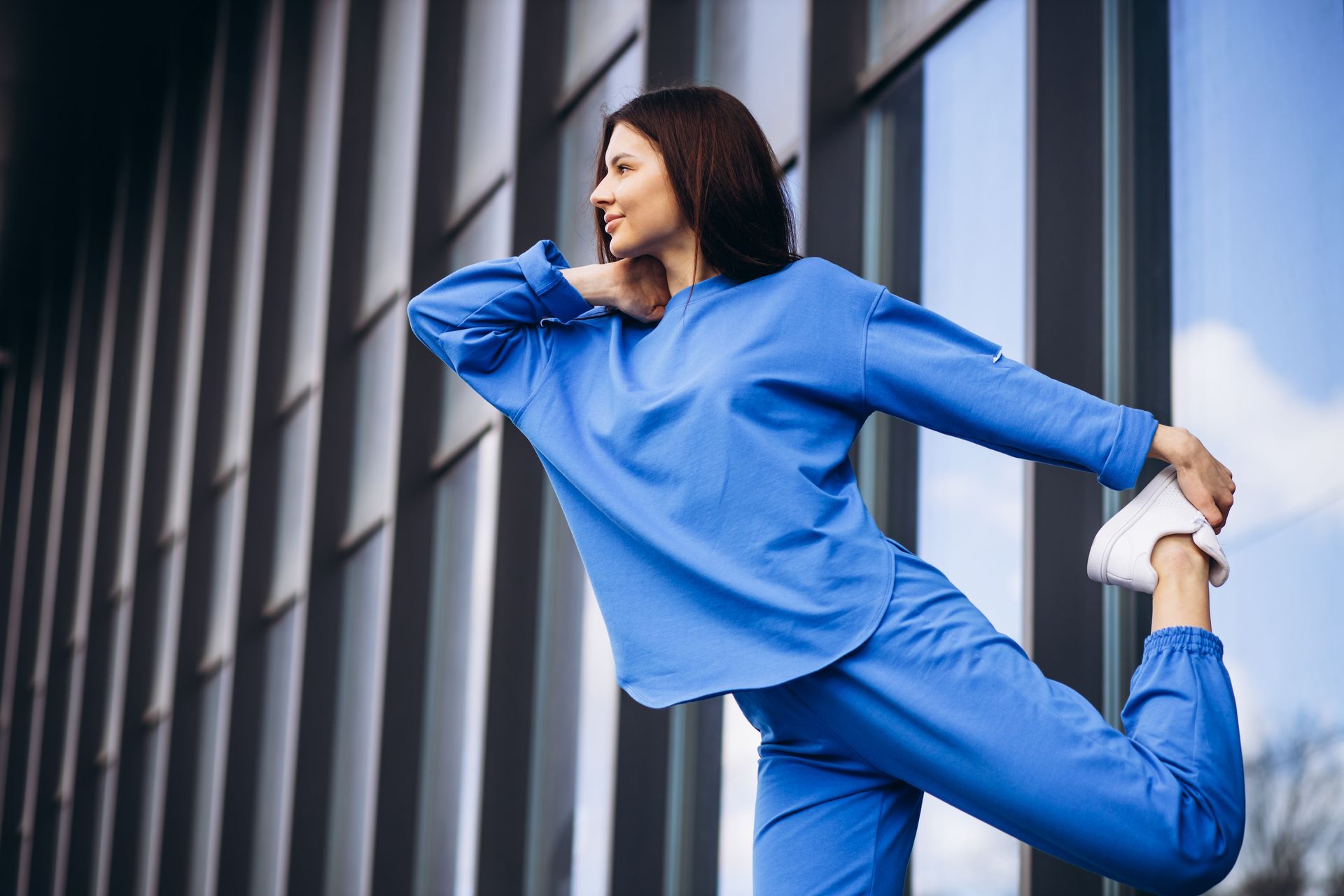 A woman in a blue outfit is stretching her leg in front of a building.