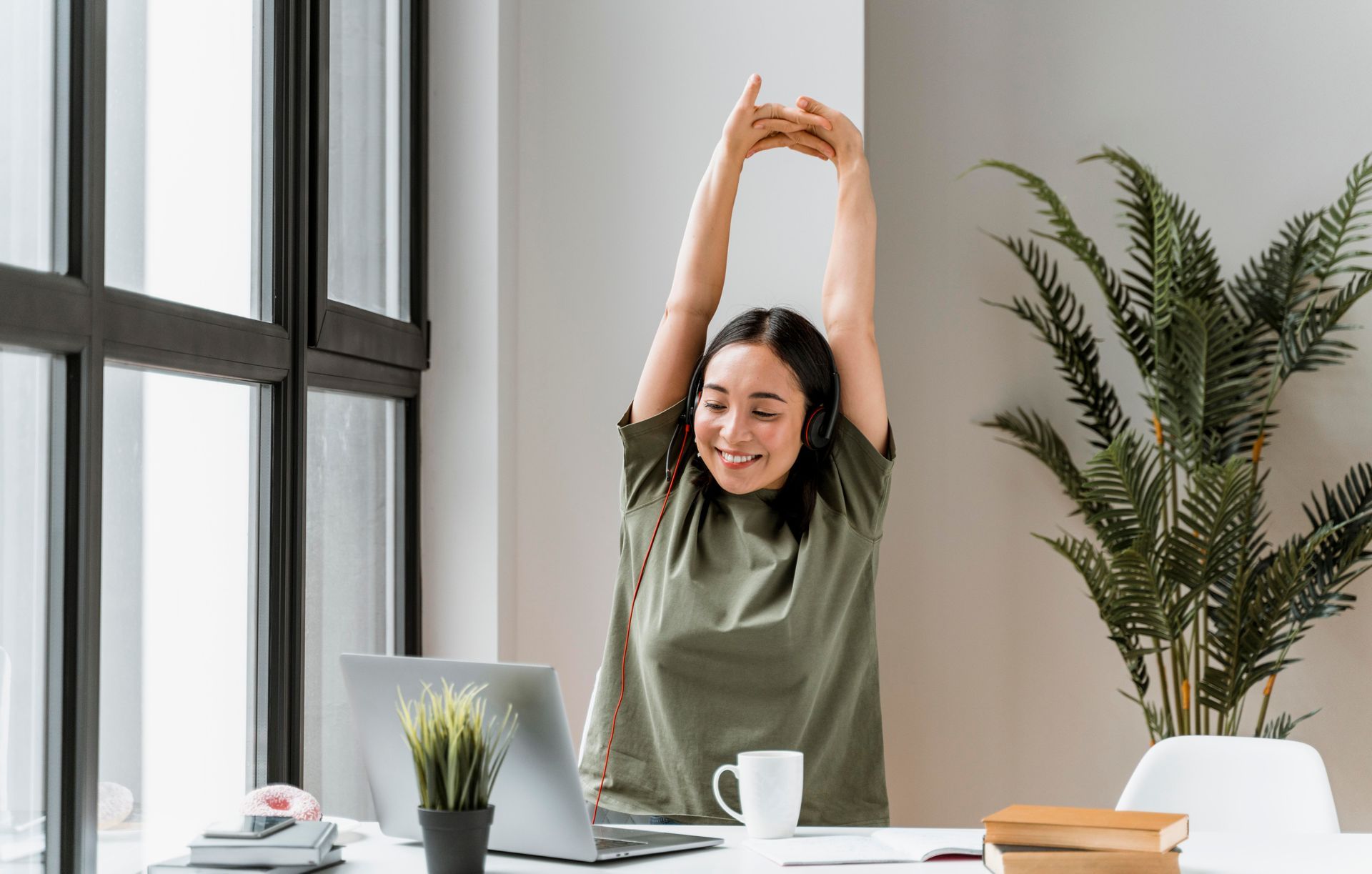 A woman is stretching her arms while sitting at a desk in front of a laptop computer.