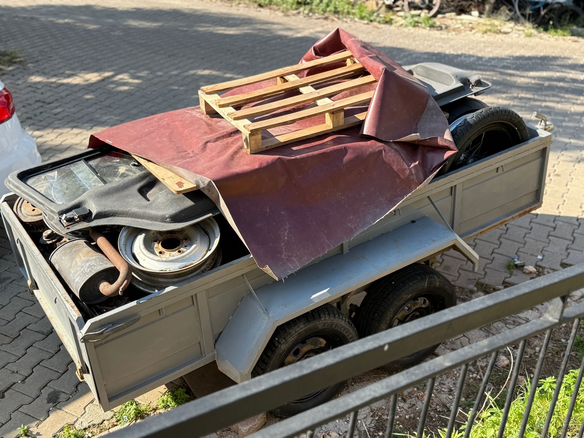 Gray trailer filled with items, covered by a maroon tarp and topped with a wooden pallet. Parked outdoors. Gray trailer filled with items, covered by a maroon tarp and topped with a wooden pallet. Parked outdoors.