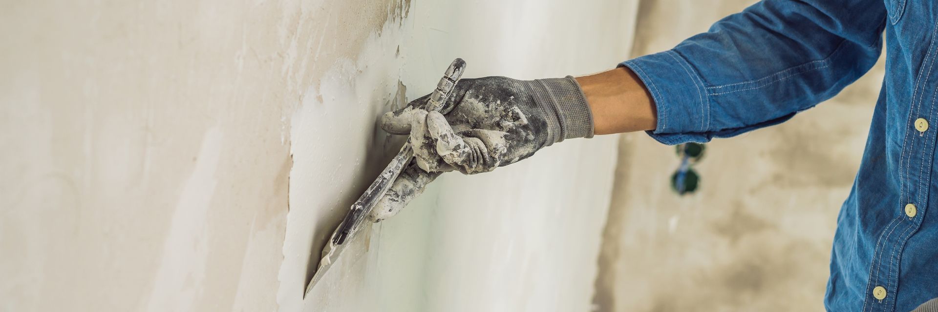 Person applying plaster to a wall with a trowel, wearing a blue shirt and work gloves. Person applying plaster to a wall with a trowel, wearing a blue shirt and work gloves.