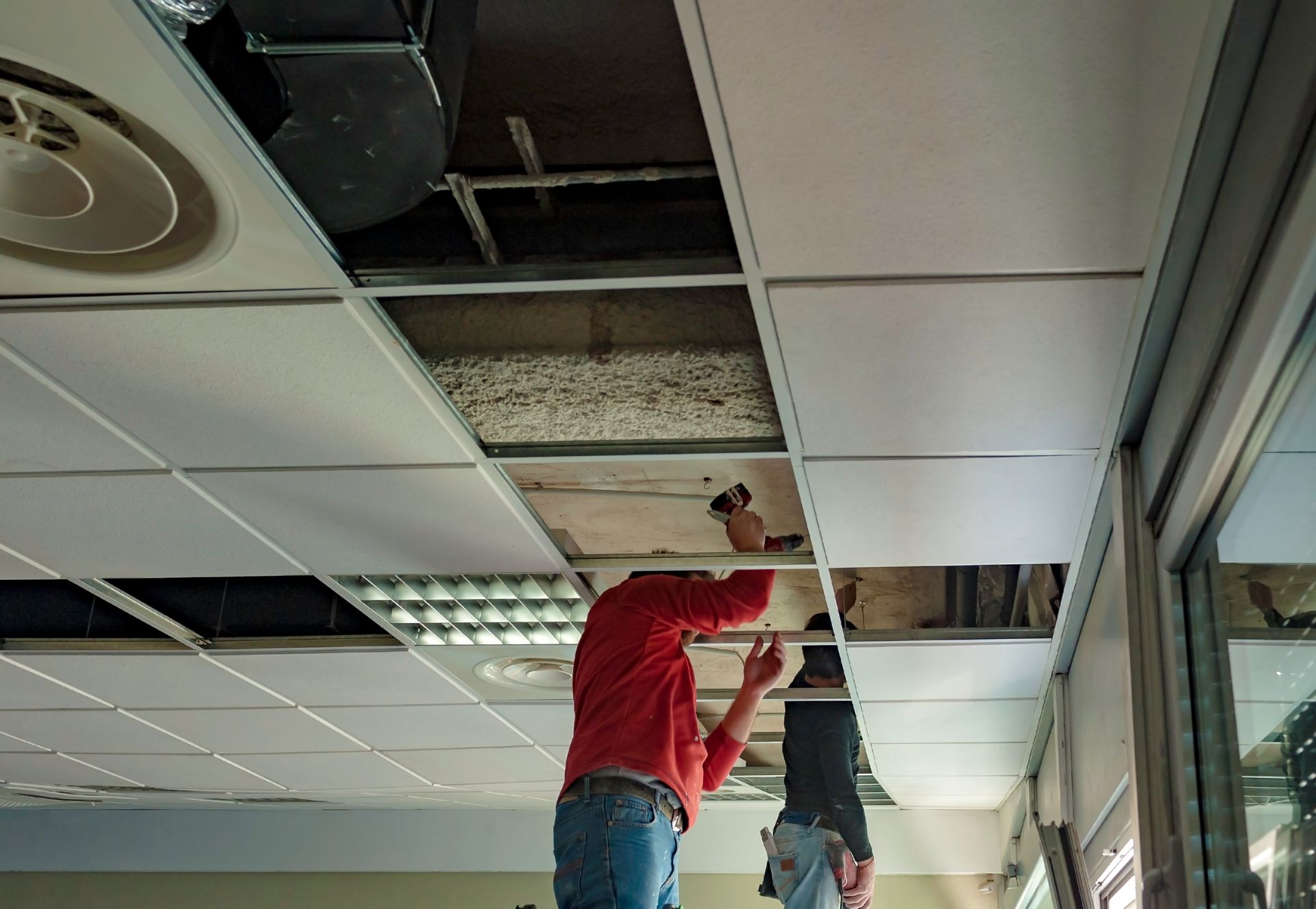 Two people installing ceiling tiles in a building, one on a ladder. Two people installing ceiling tiles in a building, one on a ladder.