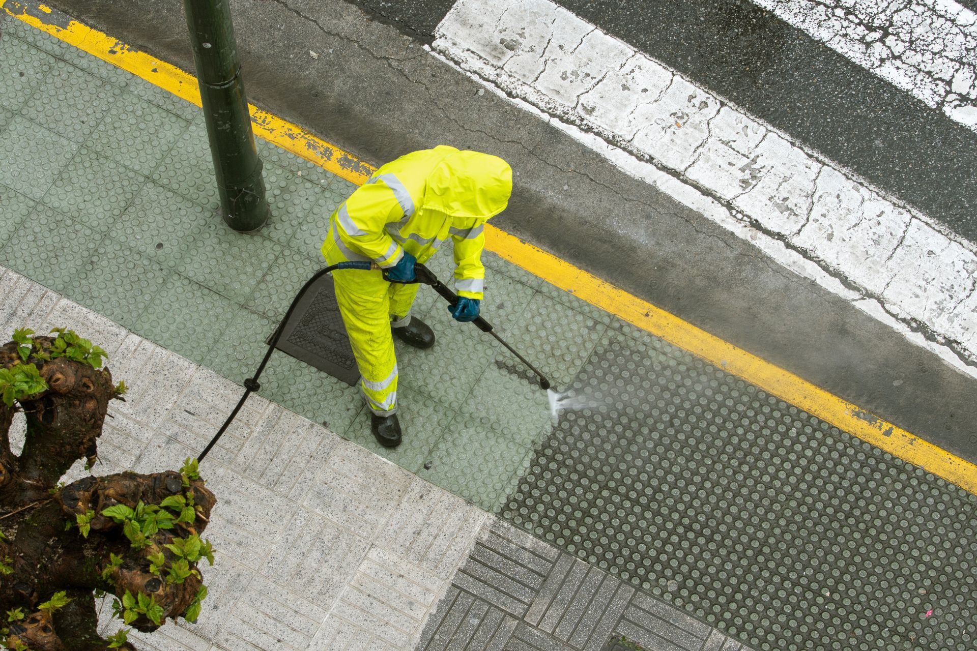Person in yellow protective suit sprays a sidewalk with a high-pressure hose.