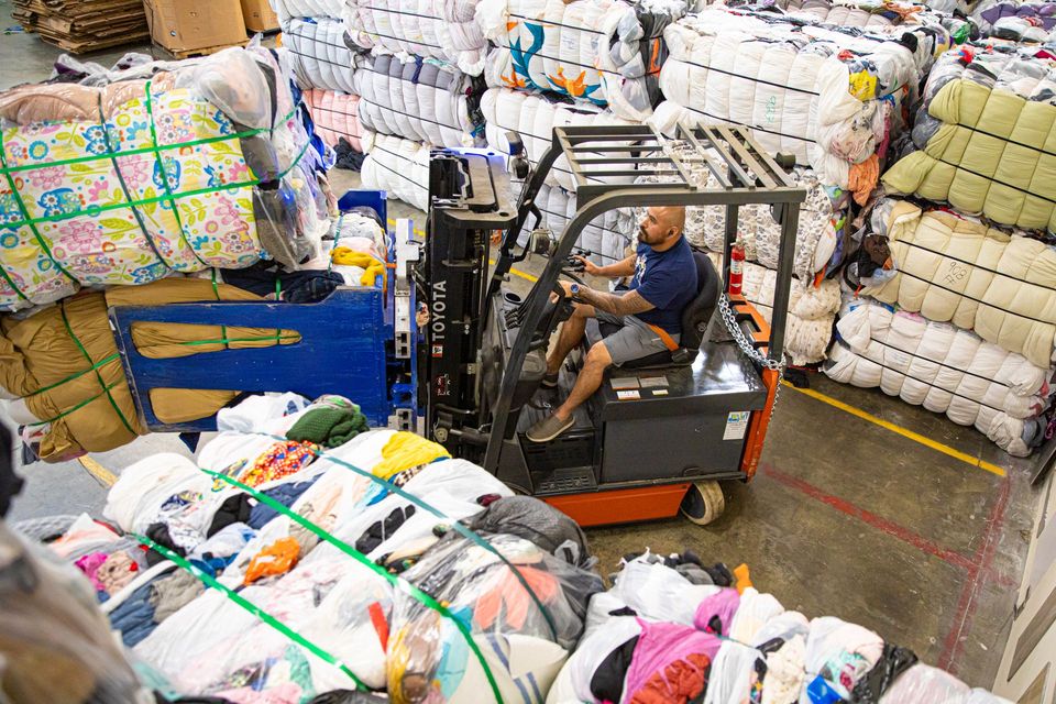 Forklift driver moving bales of textiles in a warehouse, surrounded by stacked, wrapped bundles