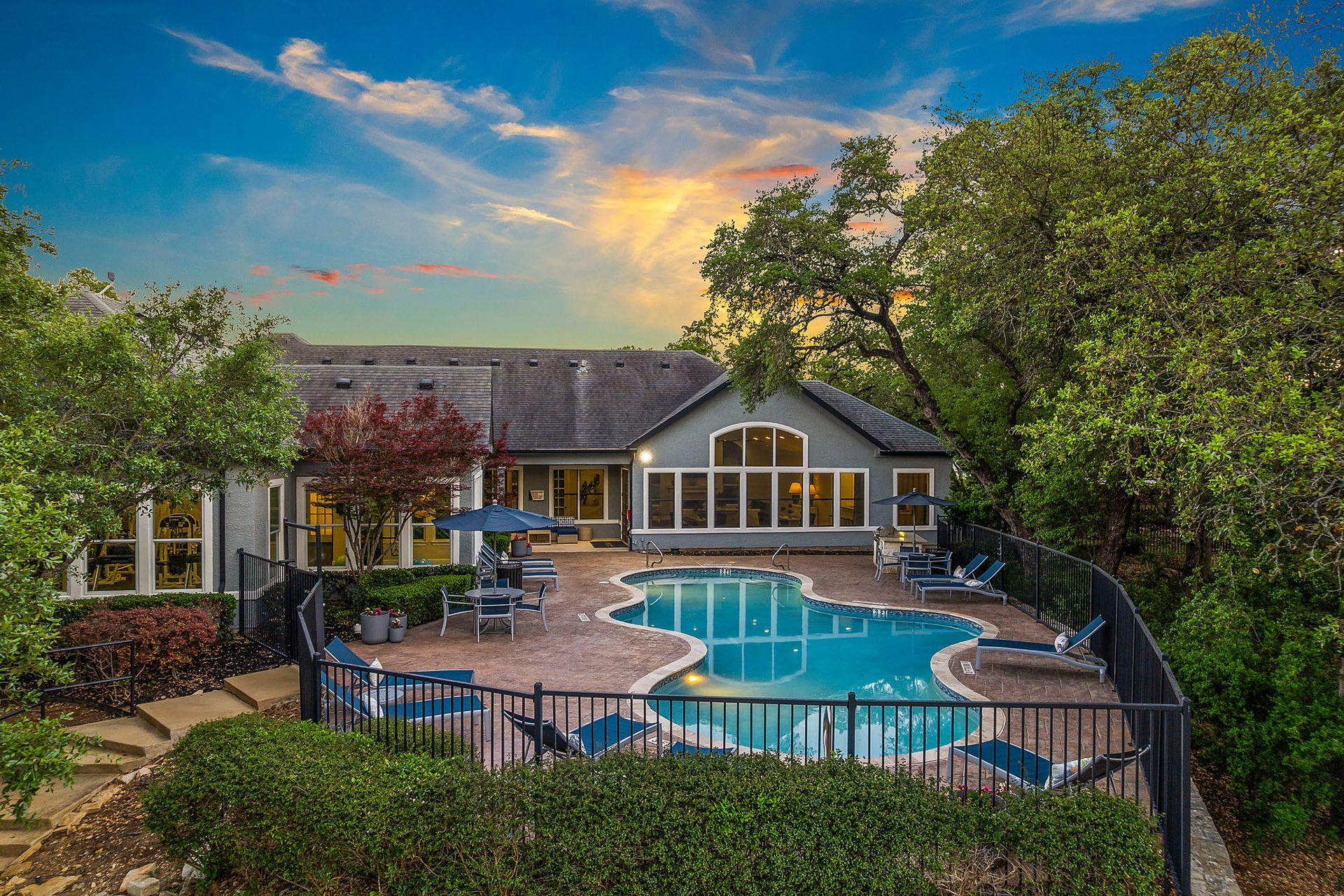 Swimming pool surrounded by a brick patio and a black fence, adjacent to a gray building with many windows and a sloped roof, under a sunset sky at Marquis at TPC, offers apartments in San Antonio, TX.