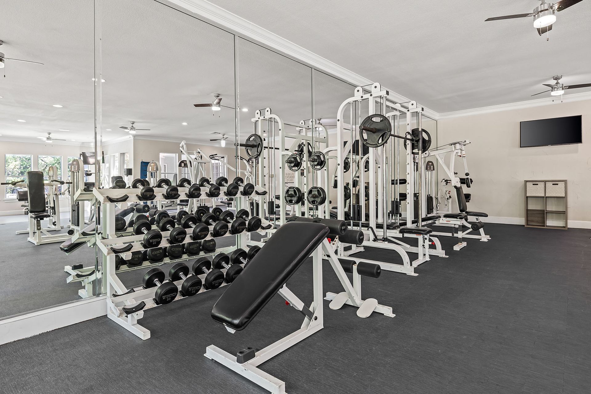 A well-equipped gym with weight machines, free weights, and an adjustable bench at Marquis at TPC, offers San Antonio apartments for rent. The room has mirrors, a TV, and dark flooring.