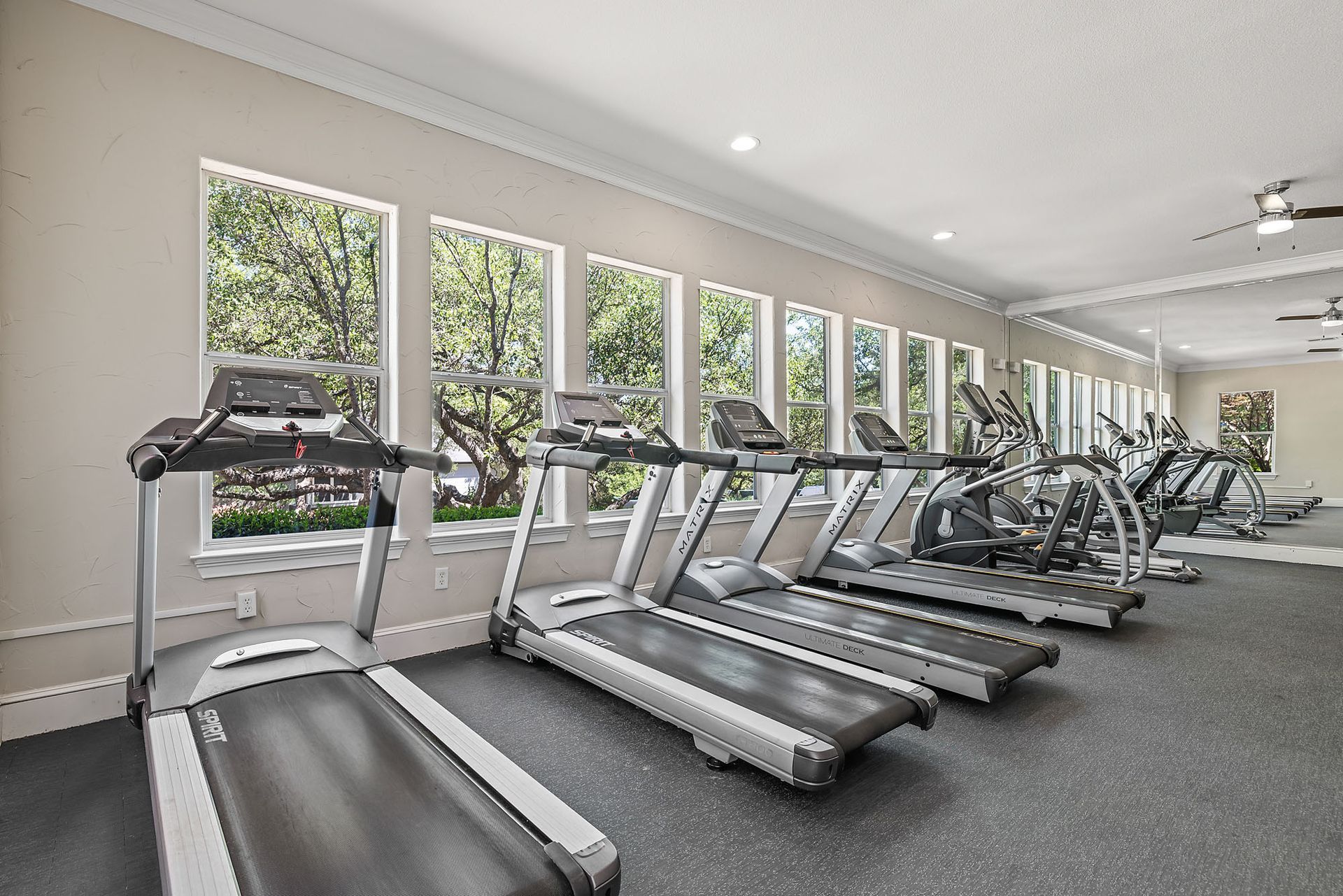 A well-lit gym with treadmills and elliptical machines lined up in front of large windows at Marquis at TPC, offers townhomes in San Antonio, TX.