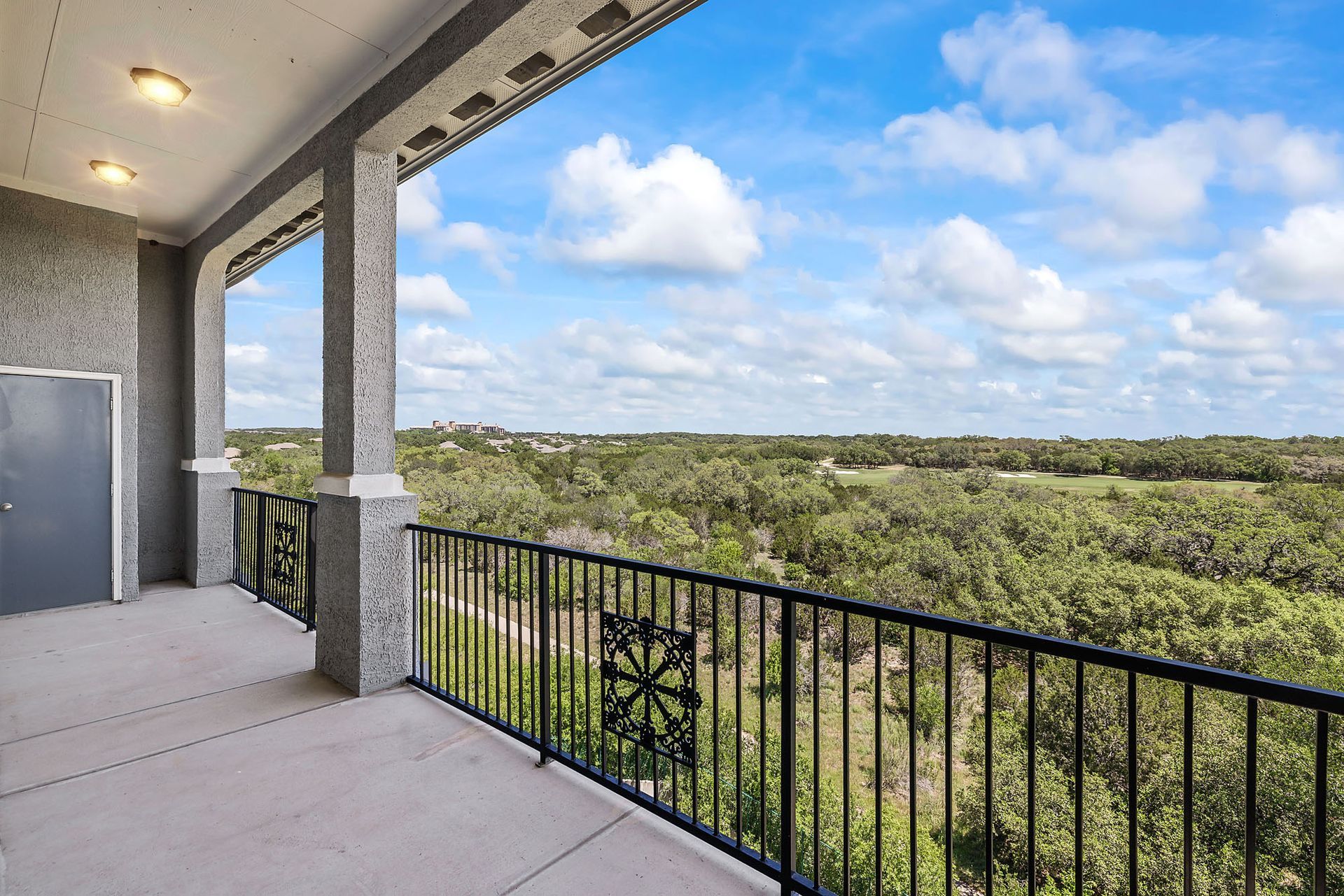 Balcony overlooking a vast green landscape under a partly cloudy sky at Marquis at TPC, offers pet-friendly apartments in San Antonio, TX. Black railing, gray columns, and a dark door are visible.