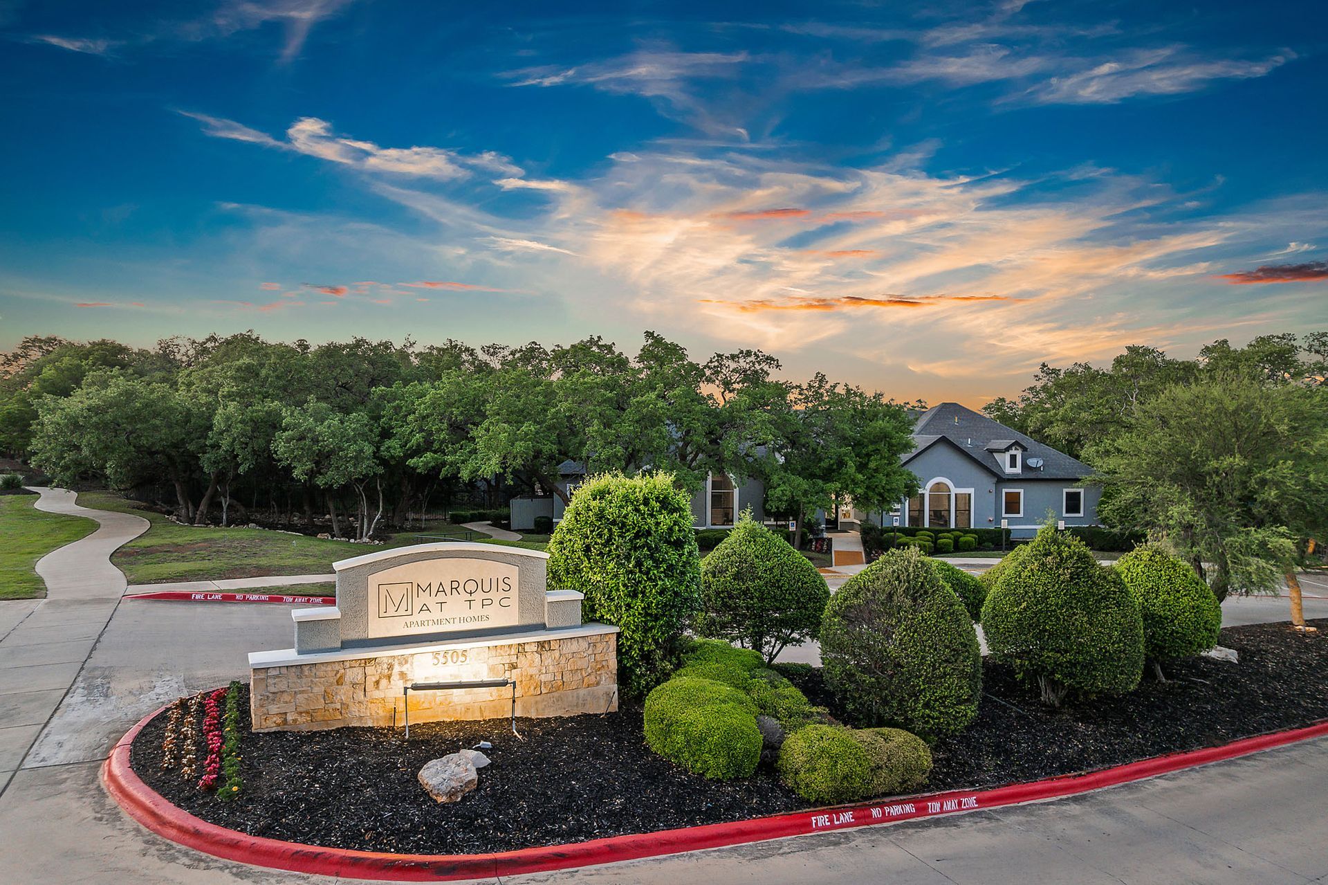 Entrance sign for a residential neighborhood with manicured bushes and a blue house visible in the background under a sunset sky at Marquis at TPC, offers apartments for rent in San Antonio, TX.