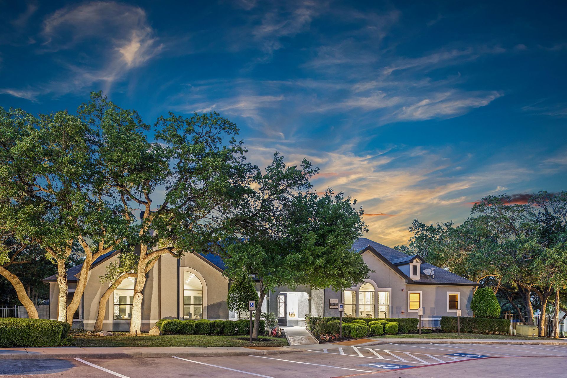 A gray building with arched windows and a dark roof under a colorful sunset sky, framed by green trees at Marquis at TPC, offers pet-friendly apartments in San Antonio, TX.