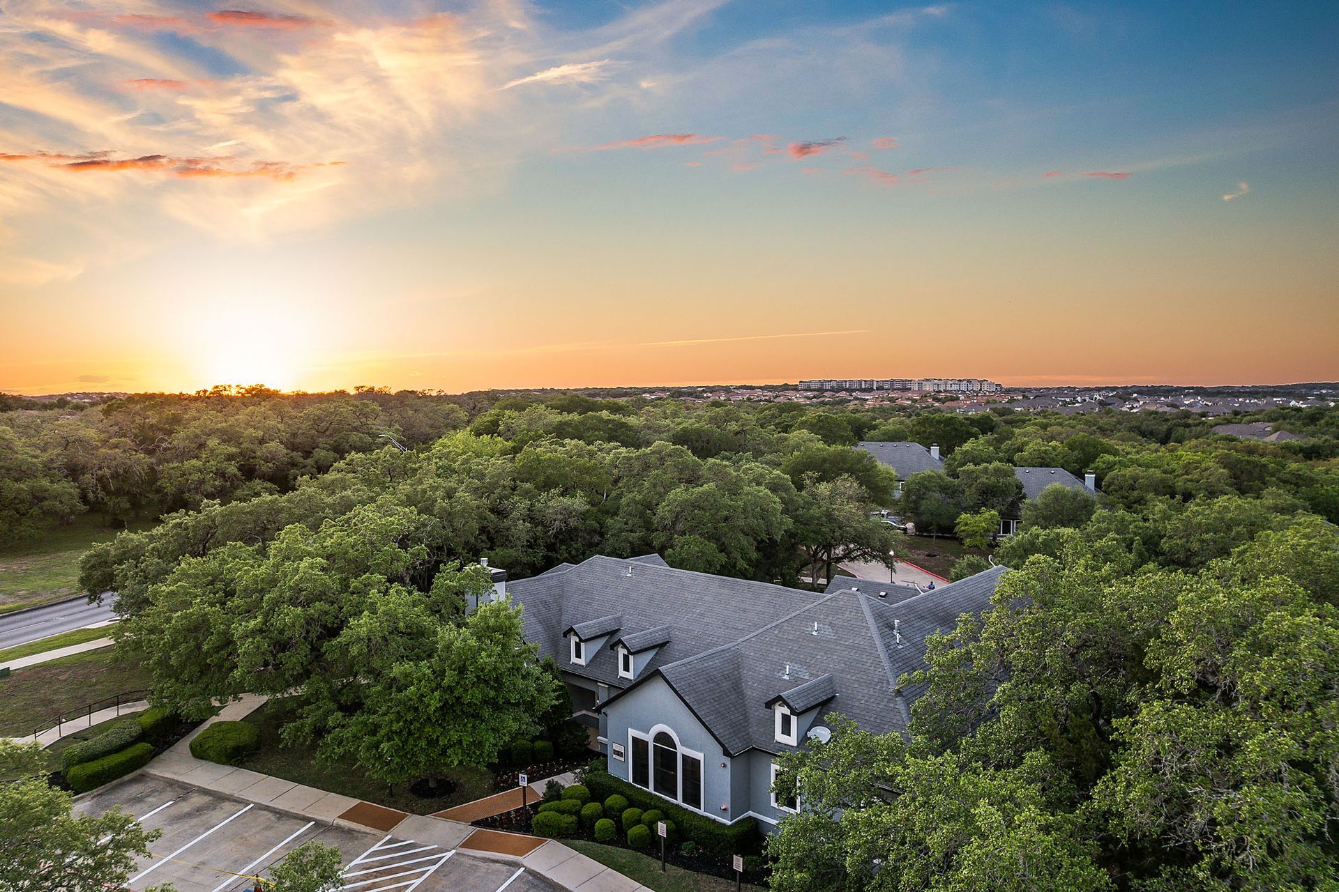 Aerial view of a building with a gray roof surrounded by green trees at sunset at Marquis at TPC, offers townhomes in San Antonio, TX. The sky is filled with orange and blue hues.
