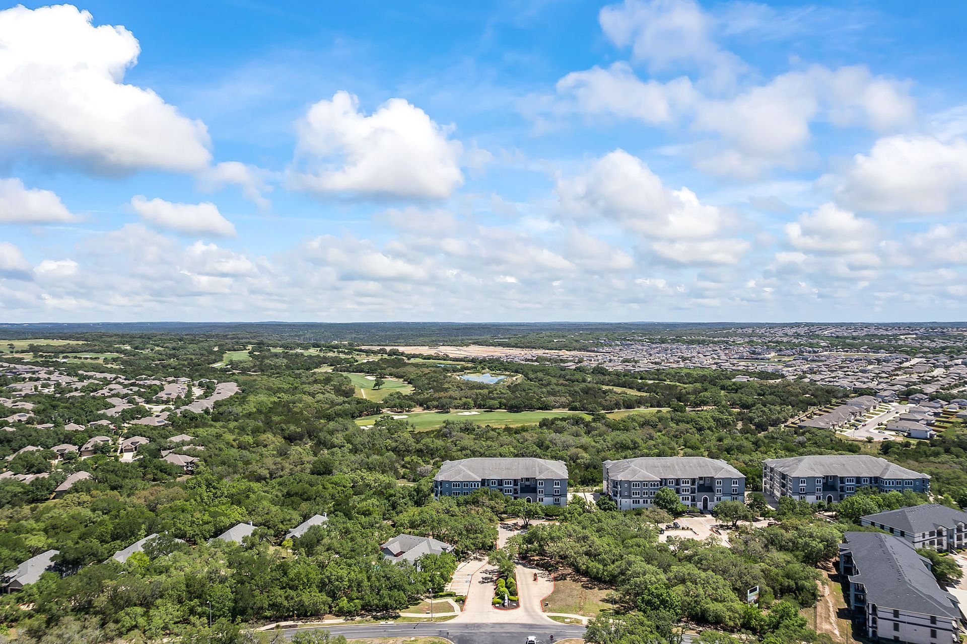 Aerial view of a residential area with apartment buildings surrounded by lush green trees, under a partly cloudy blue sky at Marquis at TPC, offers pet-friendly townhomes in San Antonio, TX.