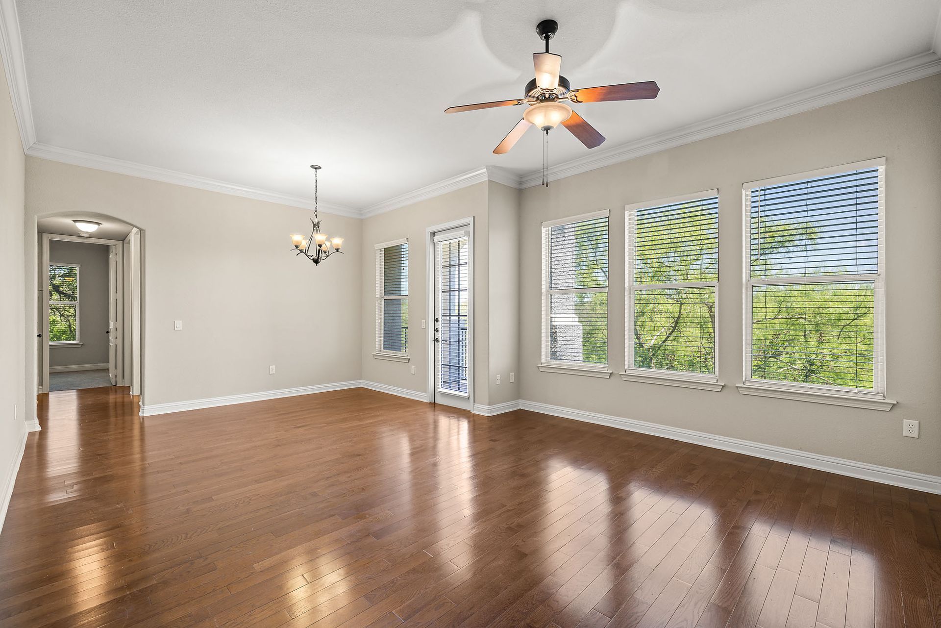 Empty living room with hardwood floors, crown molding, and large windows overlooking greenery at Marquis at TPC, offers apartments for rent in San Antonio, TX. A ceiling fan and chandelier are visible.