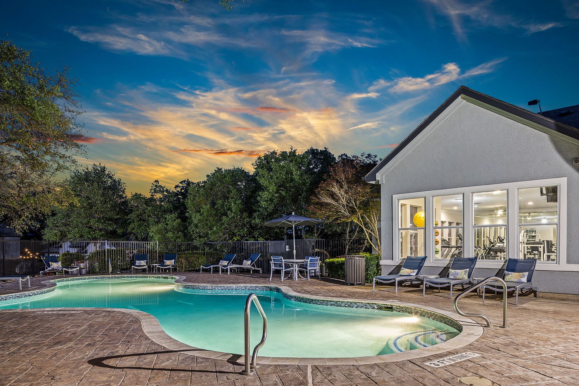 Evening view of a swimming pool and building with a gym visible through the windows at Marquis at TPC, offers apartments in San Antonio, TX. The sky is colorful with sunset hues.