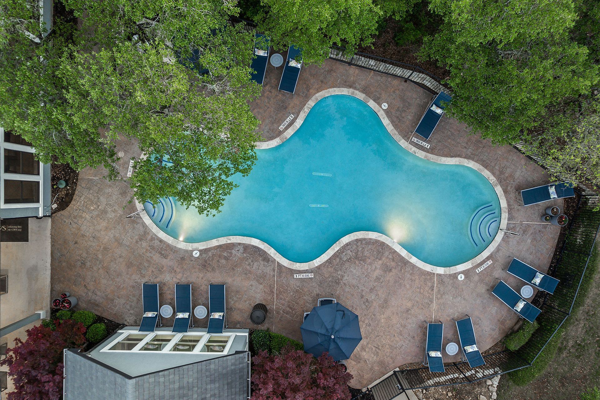 Overhead view of a freeform swimming pool surrounded by lounge chairs, trees, and a building at Marquis at TPC, offers apartments for rent in San Antonio, TX. Blue water and blue and tan color scheme.