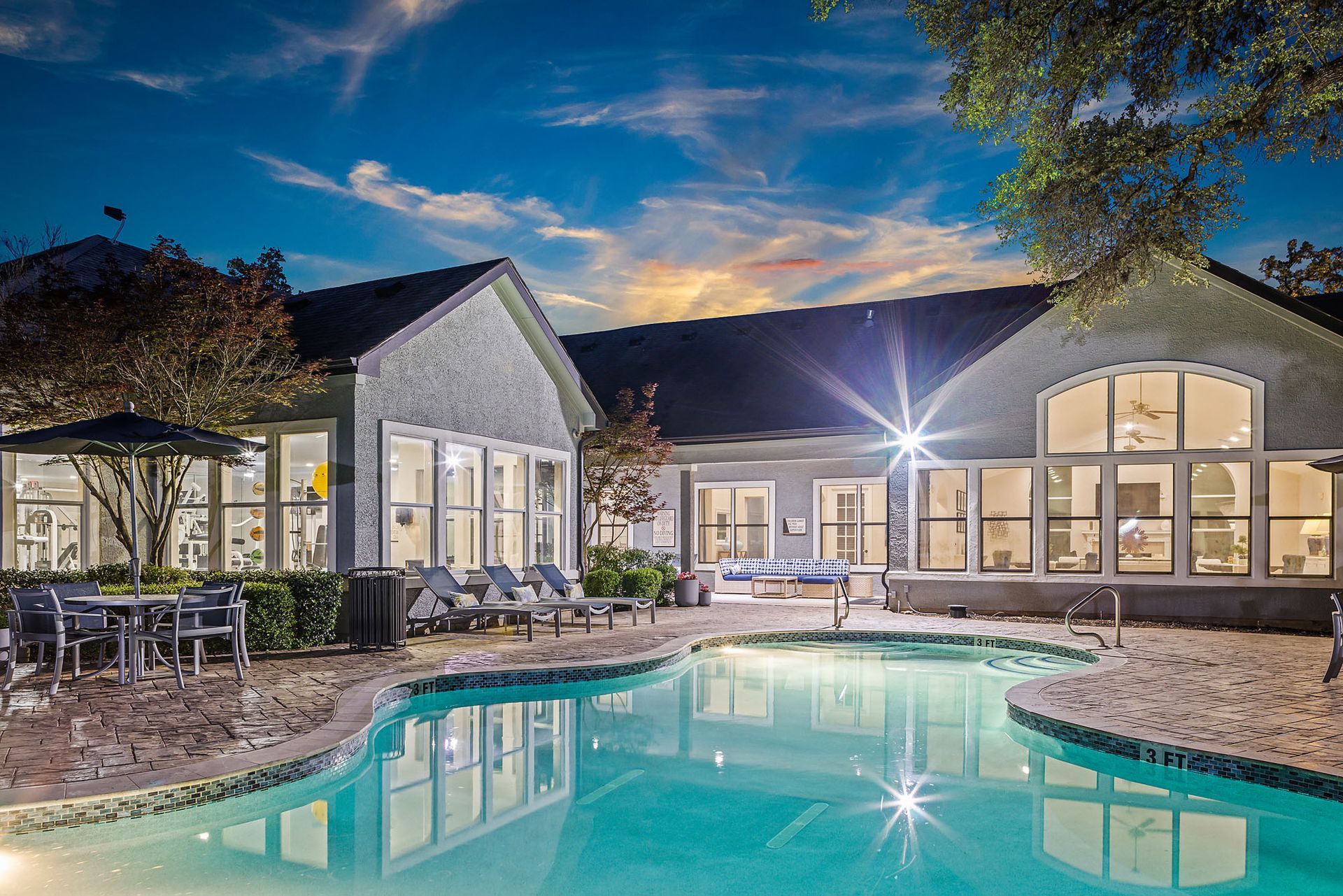 Nighttime shot of a pool in front of a building with large windows, lounge chairs, and tables at Marquis at TPC, offers apartments for rent in San Antonio, TX. The sky is blue and orange, suggesting sunset.