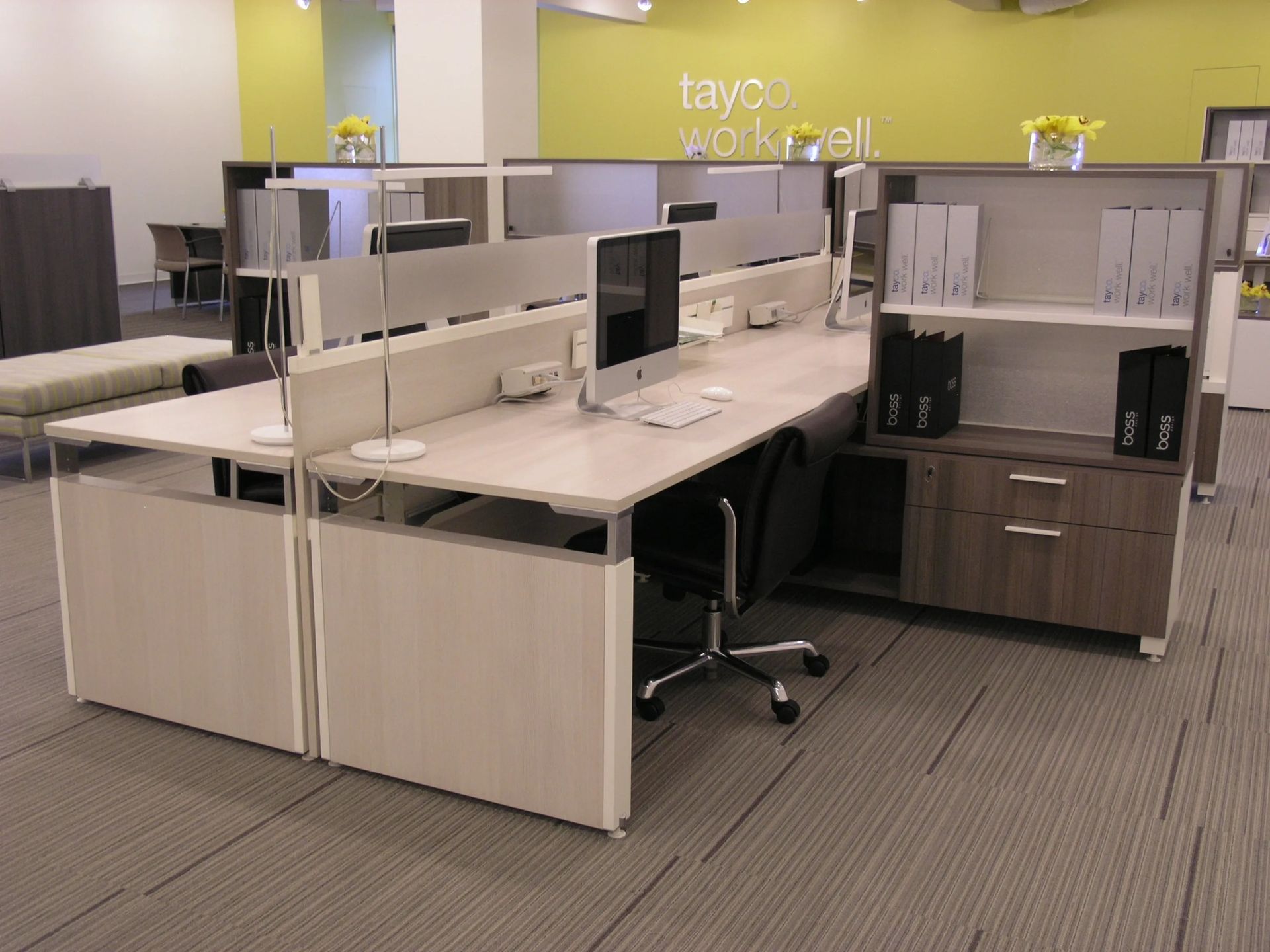 Open office cubicles with desks, monitors, and chairs in a light wood and gray workspace.