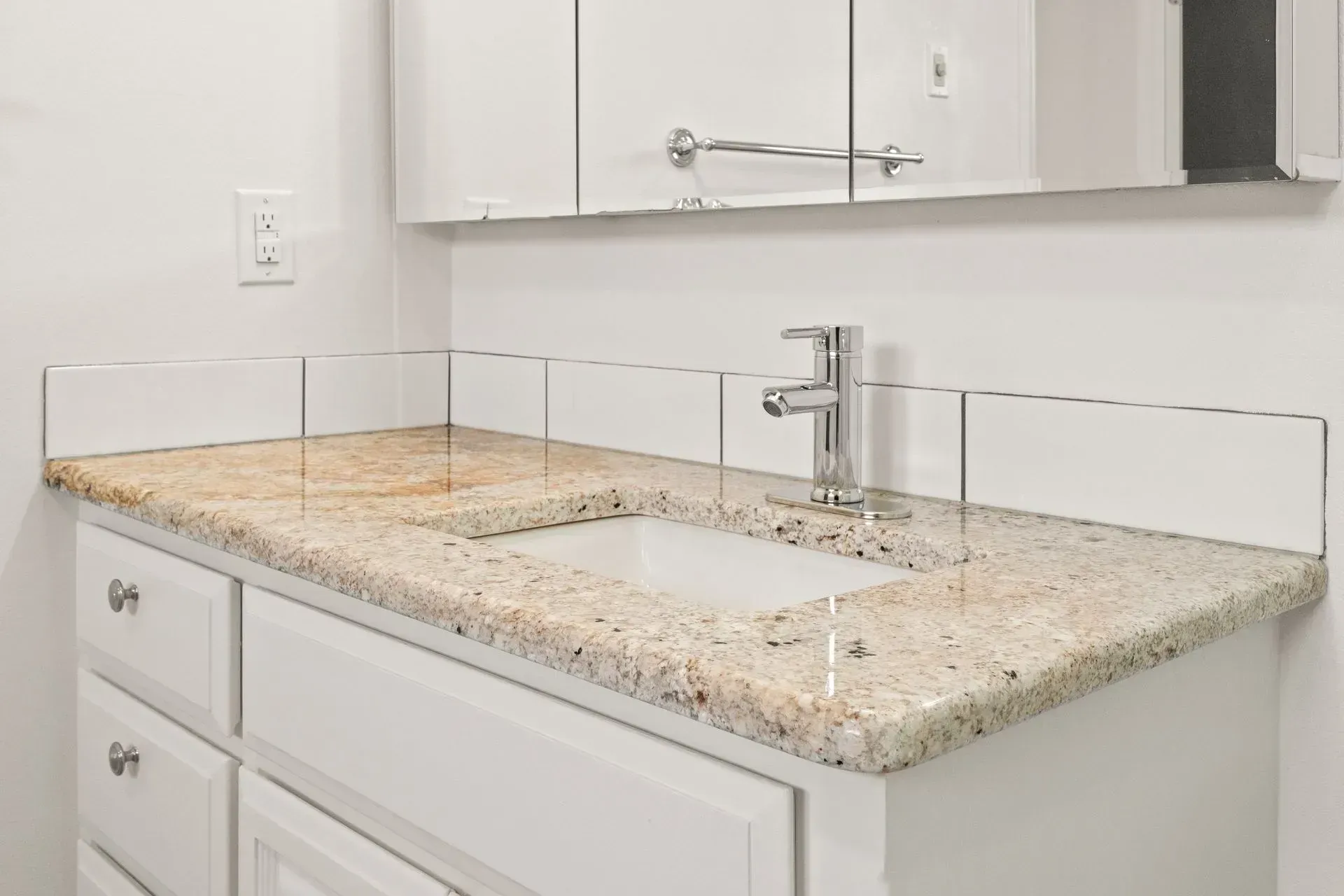 Bathroom vanity with white cabinets, beige granite countertop, and white tile backsplash.
