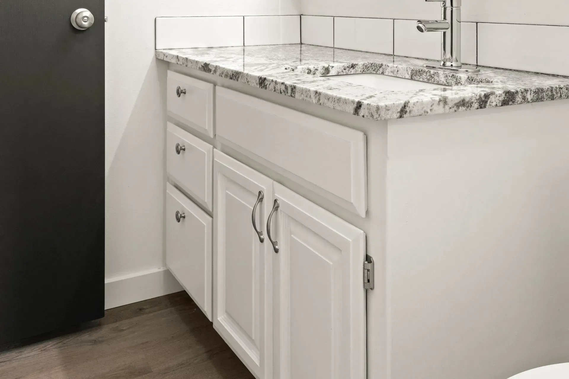 White bathroom vanity with granite countertop, cabinets, and sink. Silver hardware and faucet. Dark door on left.
