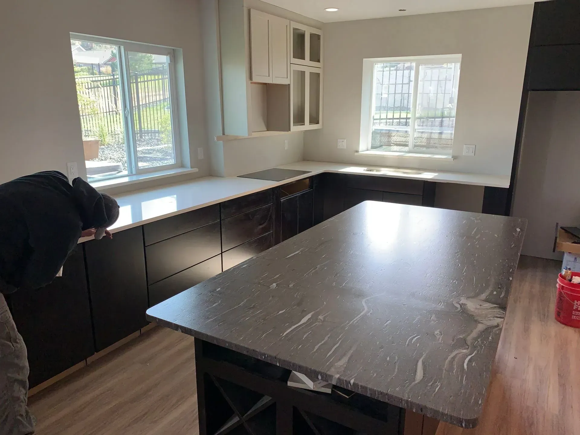 Kitchen with dark cabinets, island, white countertops, two windows, a person leans over counter.