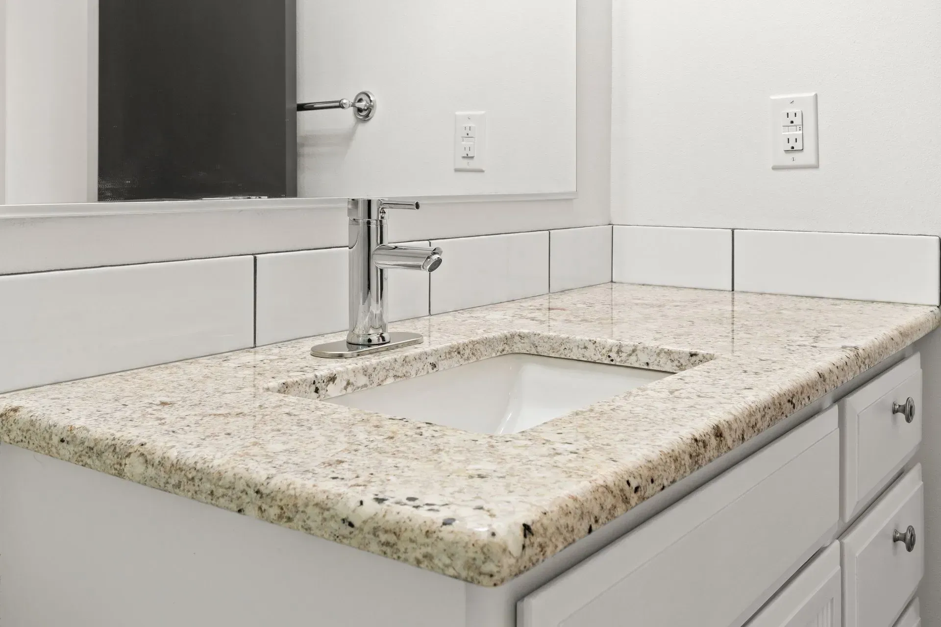 Bathroom vanity with granite countertop, white sink, and white cabinets against a white wall.