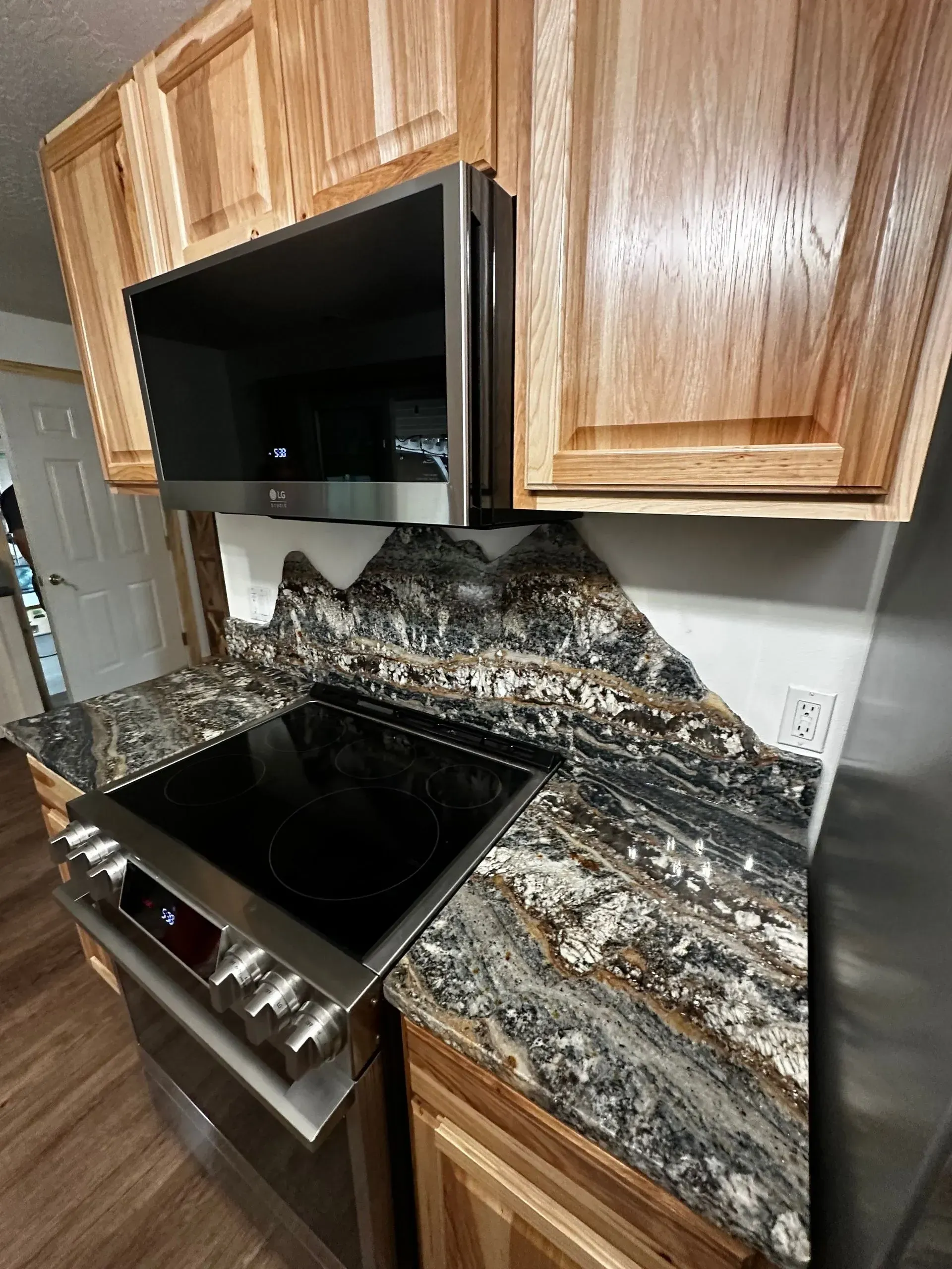 Kitchen with light wood cabinets, dark countertop, and stainless steel appliances, including a cooktop and microwave.