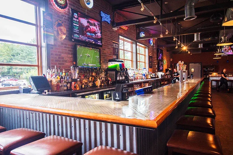 A long bar in a restaurant with stools and televisions on the wall.