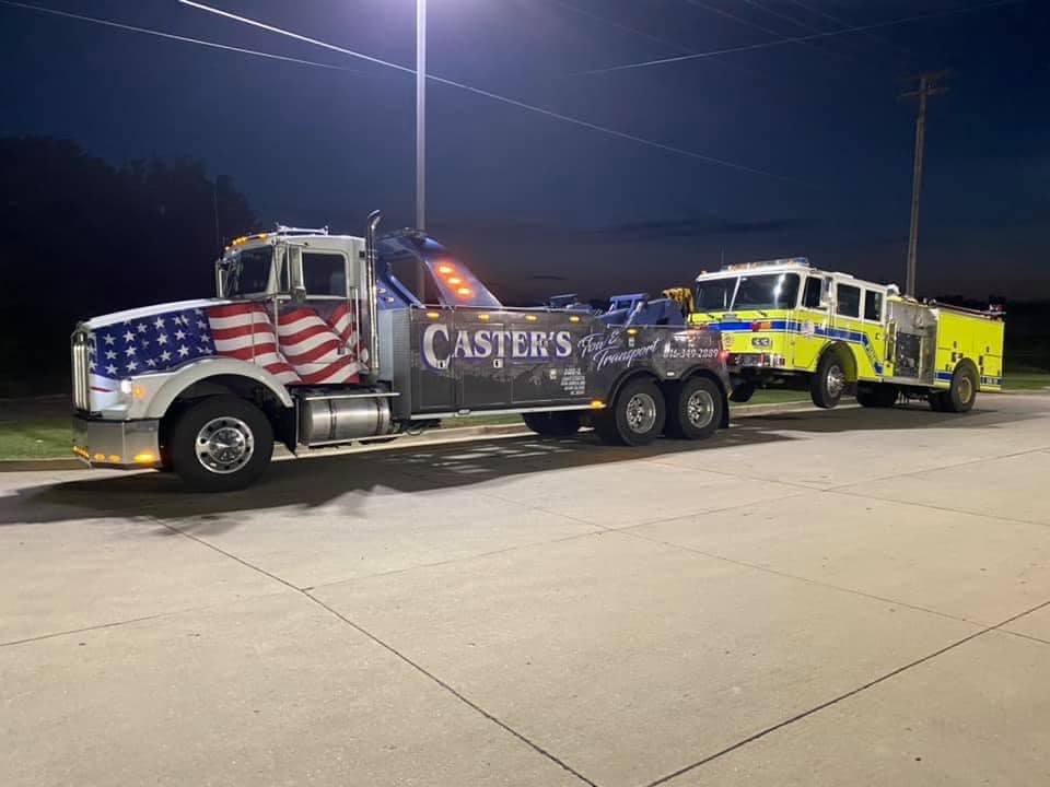 A tow truck with a patriotic flag design is towing a yellow fire truck at night. 