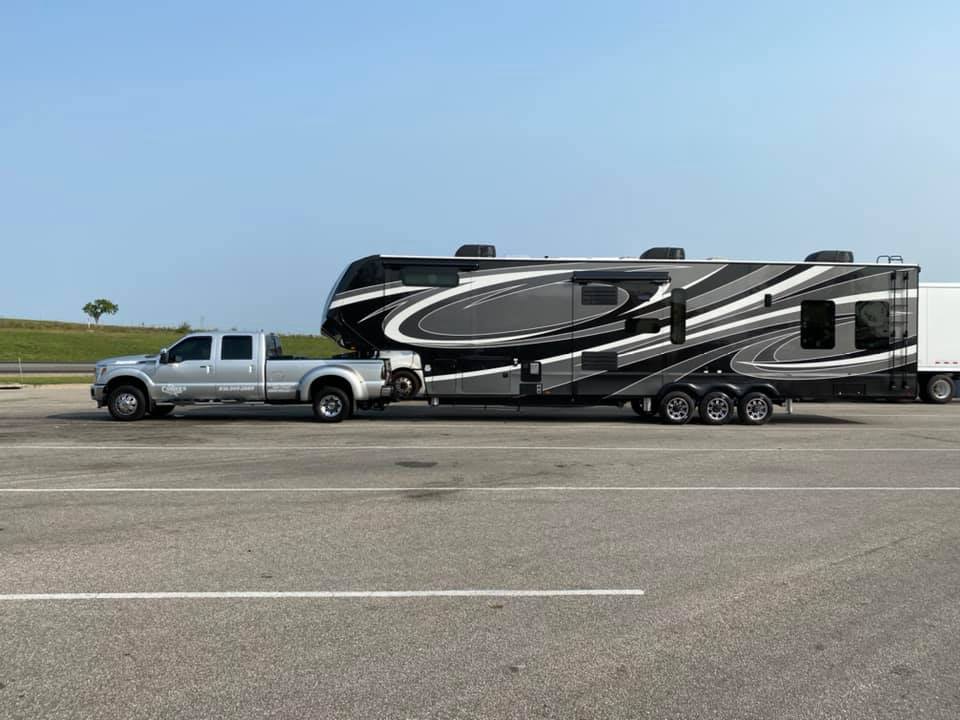 Silver pickup truck towing a large, black and gray fifth-wheel RV in a parking area under a blue sky.