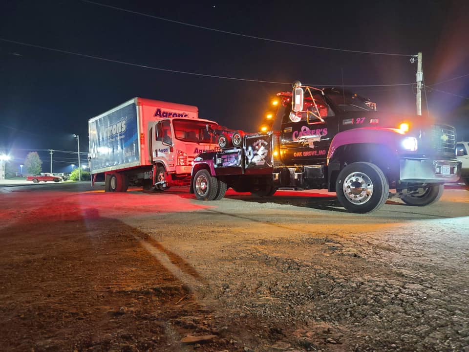 A tow truck pulling a box truck at night on a dark road, illuminated by the tow truck's lights.