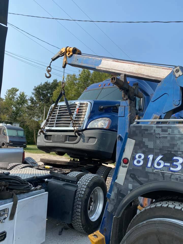 A blue tow truck lifting a blue semi-truck cab. The setting is outdoors on a sunny day.