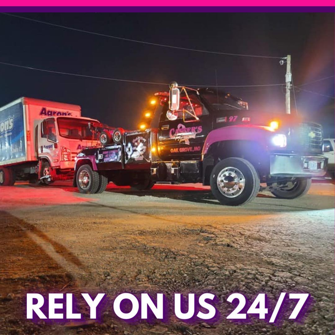 A tow truck pulling a moving truck at night. The tow truck is purple and has bright lights. Text reads 