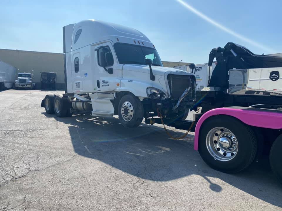 A white semi-truck being towed by another truck, both parked in a lot on a sunny day. The semi has front-end damage.
