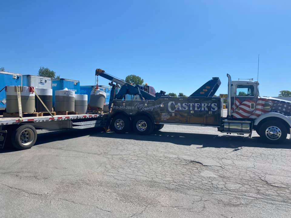 A tow truck loading cylindrical containers onto a flatbed trailer under a blue sky. The truck has 