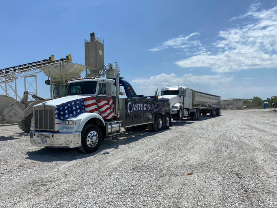 A tow truck decorated with a semi-trailer on a gravel lot in front of an industrial plant on a sunny day.