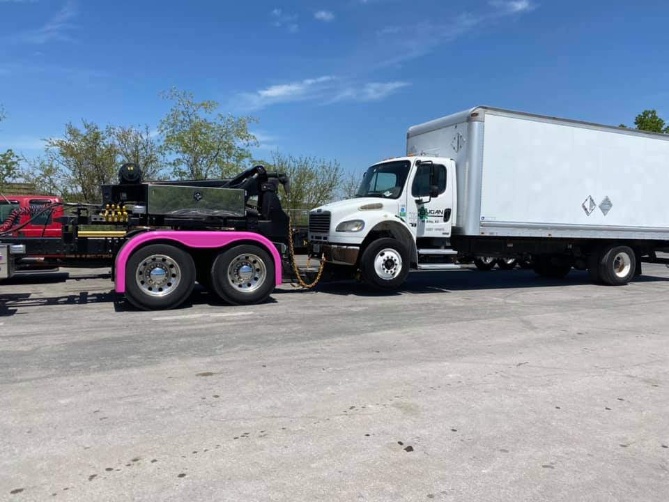 White truck with a box trailer being towed by a truck with pink fenders in a parking lot on a sunny day.