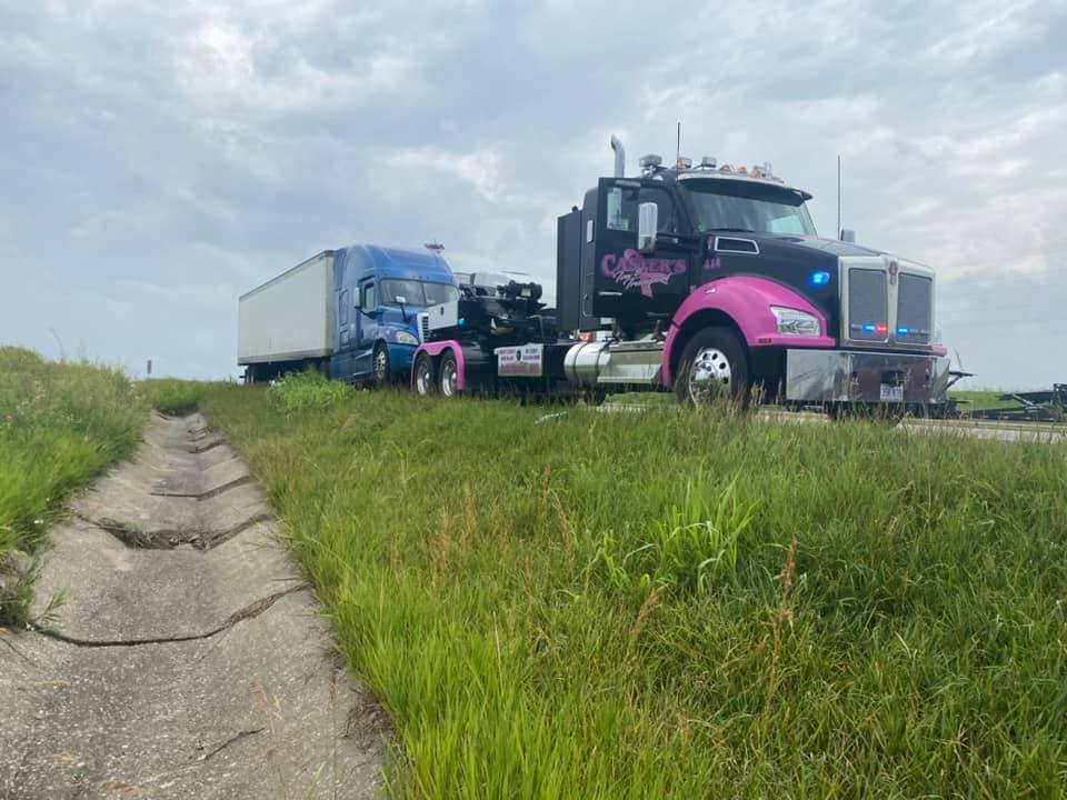 A black and pink tow truck pulls a blue semi-truck out of a grassy ditch on a cloudy day.