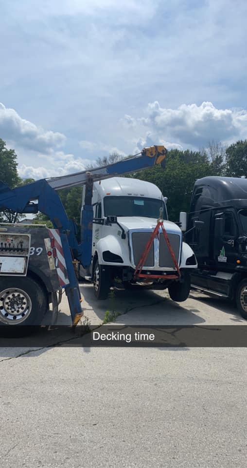 A tow truck lifting a white semi-truck with a blue crane. The setting is outdoors on a bright day.