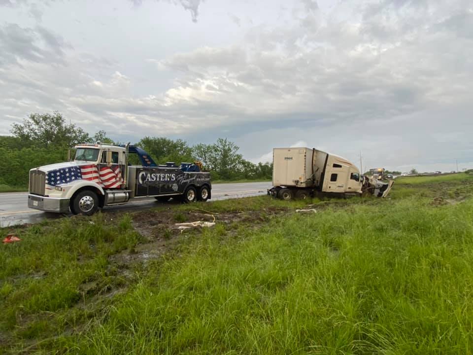 Tow truck pulling a semi-truck that is off the road in a grassy area. Cloudy sky overhead.