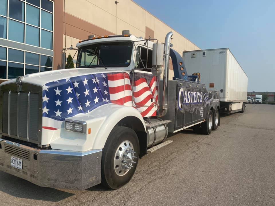 Tow truck with American flag design on the cab, towing a white trailer. Parked outside a large industrial building.