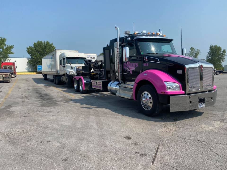 Black semi-truck with pink accents towing a smaller white truck in a parking lot on a sunny day.