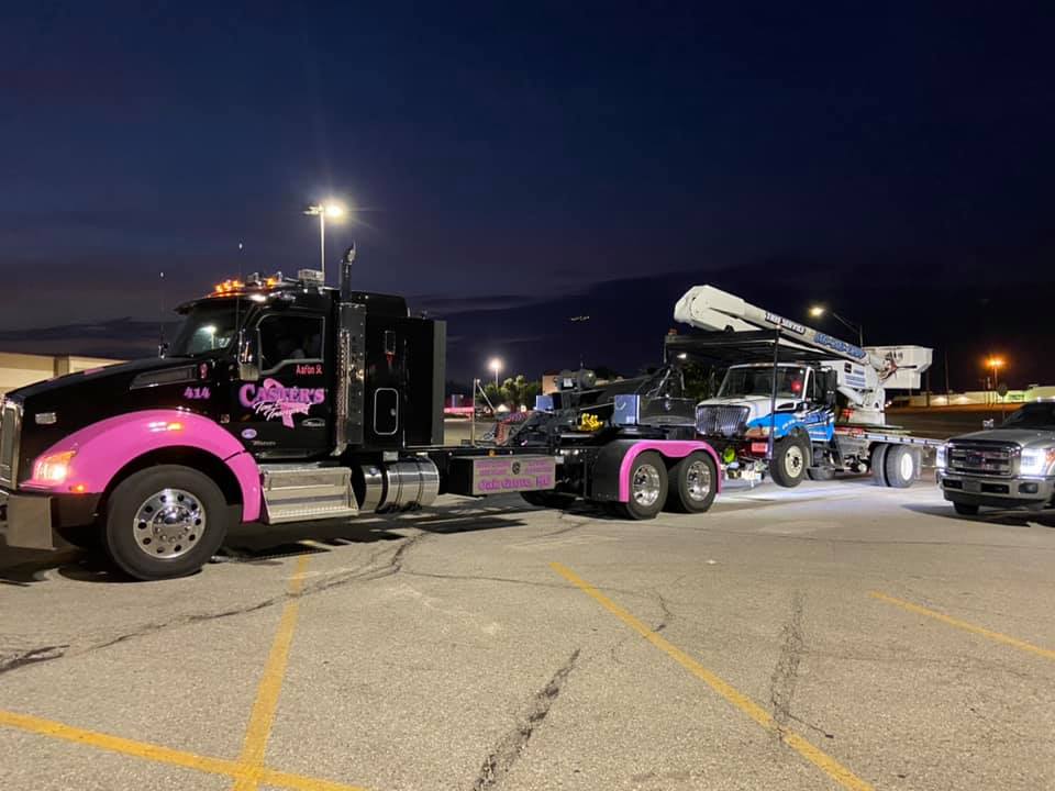 A black semi-truck with pink accents towing a utility truck on a flatbed trailer in a parking lot at dusk.