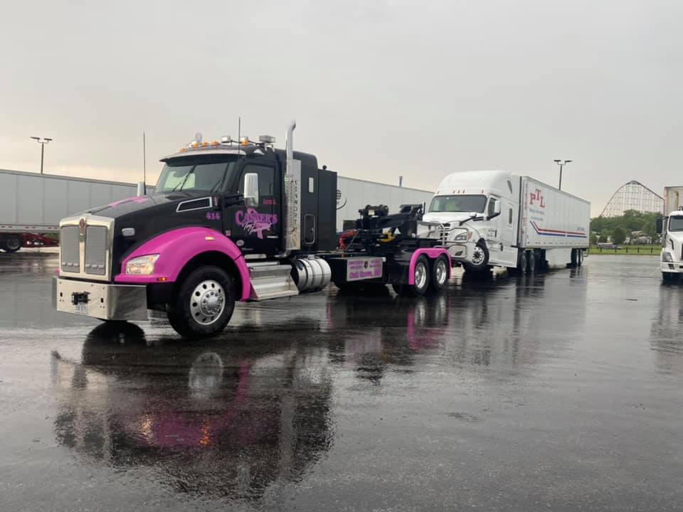 A black and pink tow truck towing a white semi-truck in a wet parking lot. 