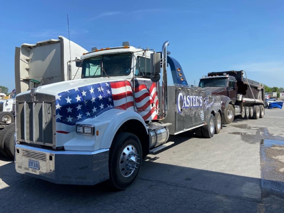 A tow truck with an American flag design on the front pulling a dump truck in a sunny parking lot.